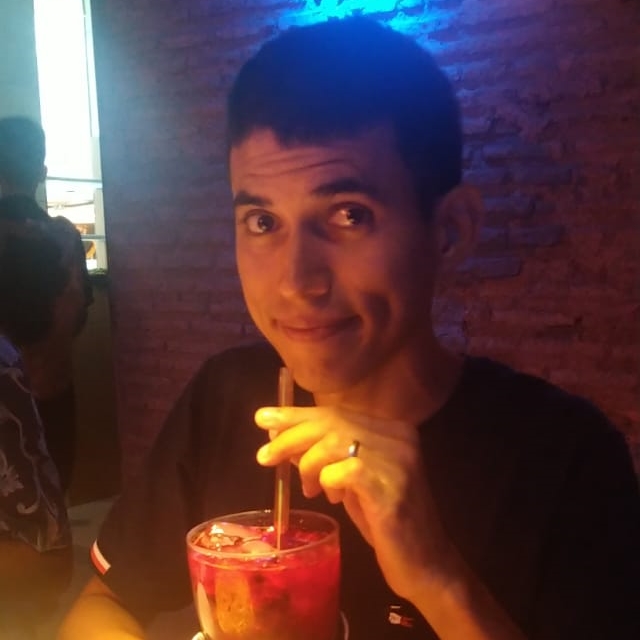 A picture of me smiling, holding a glass filled with a red beverage with ice cubes and a straw. Background is a brick wall.